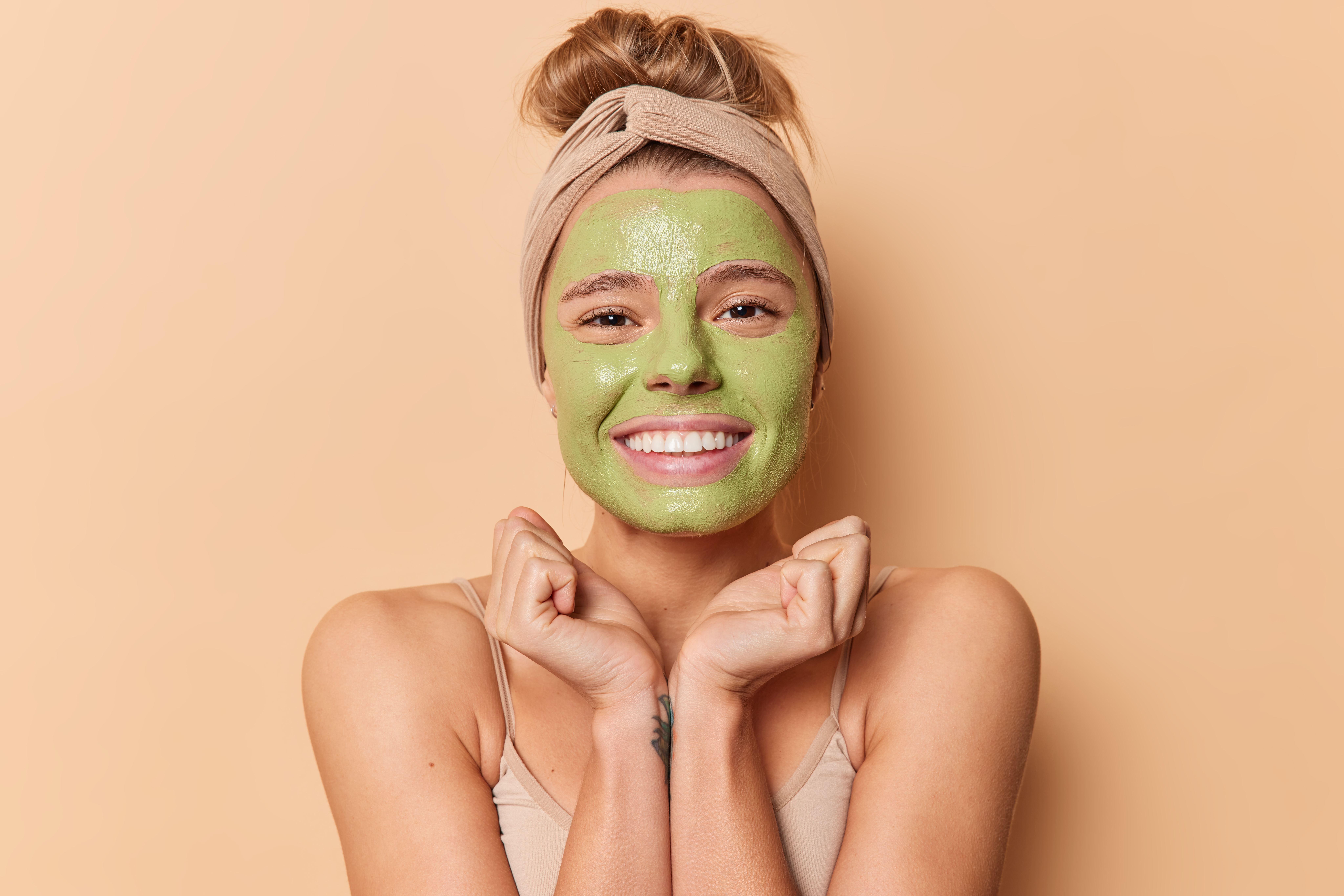 Happy young European woman keeps hands under chin smiles gladfully applies green nourishing mask wears headband undergoes beauty procedures for skin treatment isolated over beige background.